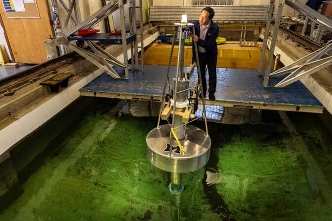 A metal buoy floats inside an indoor tank of water. A man closely inspects an illuminated light bulb attached to the top of a post on the buoy.