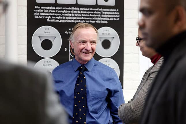 A male professor in a blue shirt and patterned block M tie smiles while interacting with a group in a scientific setting. The blurred figures of attendees surround him, and a black and white scientific poster with circular diagrams and text is visible in the background.